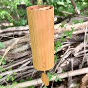 A natural Koshi River Earth Symphony Walnut wind chime hanging outdoors from a tree trunk, with a small wooden tag below and a black hanging cord, set against a forest background.