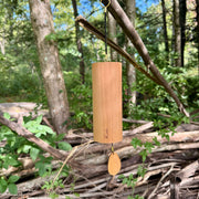 A natural Koshi River Earth Symphony  cherry wood wind chime hanging outdoors from a tree trunk, with a small wooden tag below and a black hanging cord, set against a forest background.