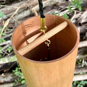A natural Koshi River Earth Symphony  cherry wood wind chime hanging outdoors from a tree trunk, with a small wooden tag below and a black hanging cord, set against a forest background.
