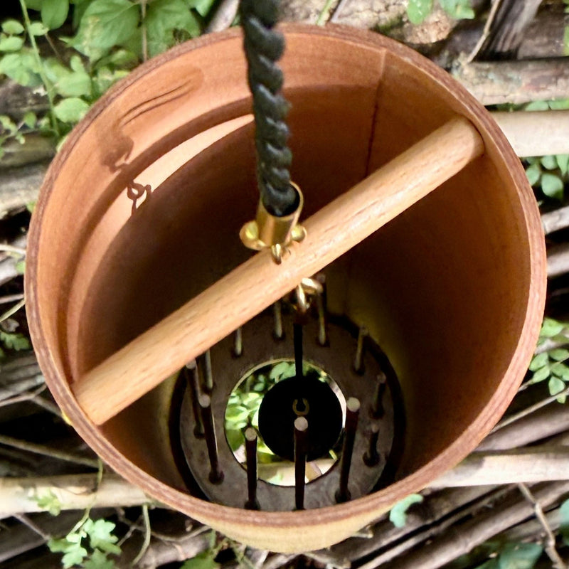the inside of A natural Koshi River Earth Symphony Cherry wind chime hanging outdoors from a tree trunk, with a small wooden tag below and a black hanging cord, set against a forest background.