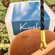 A natural Koshi River Earth Symphony  cherry wood wind chime hanging outdoors from a tree trunk, with a small wooden tag below and a black hanging cord, set against a forest background.