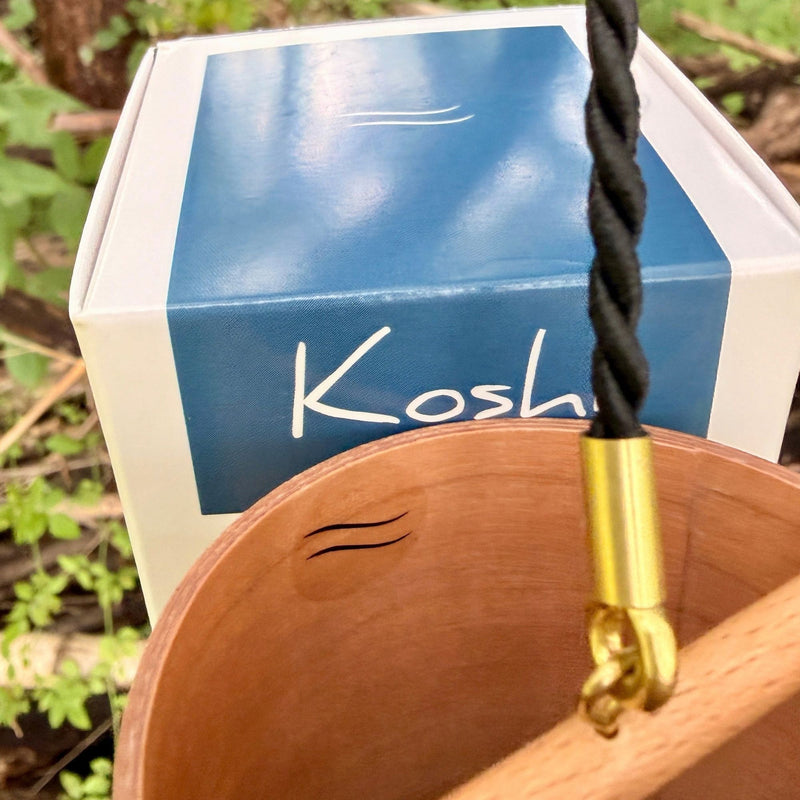 A natural Koshi River Earth Symphony  cherry wood wind chime hanging outdoors from a tree trunk, with a small wooden tag below and a black hanging cord, set against a forest background.