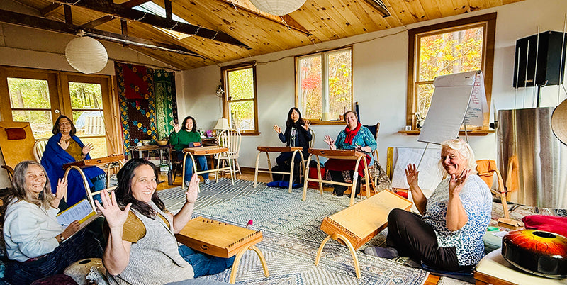 Seven women sitting in a circle with feeltone monochords in front of them waving hands to the photographer | weplaywelltogether