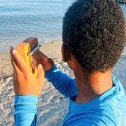 A person on a sandy beach facing the ocean, holding a wooden kalimba thumb piano.