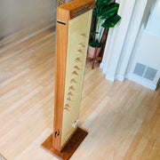 A wooden stringed instrument, likely a zither or folk guitar, standing upright on a light wood floor in an indoor room with a potted plant in the background.