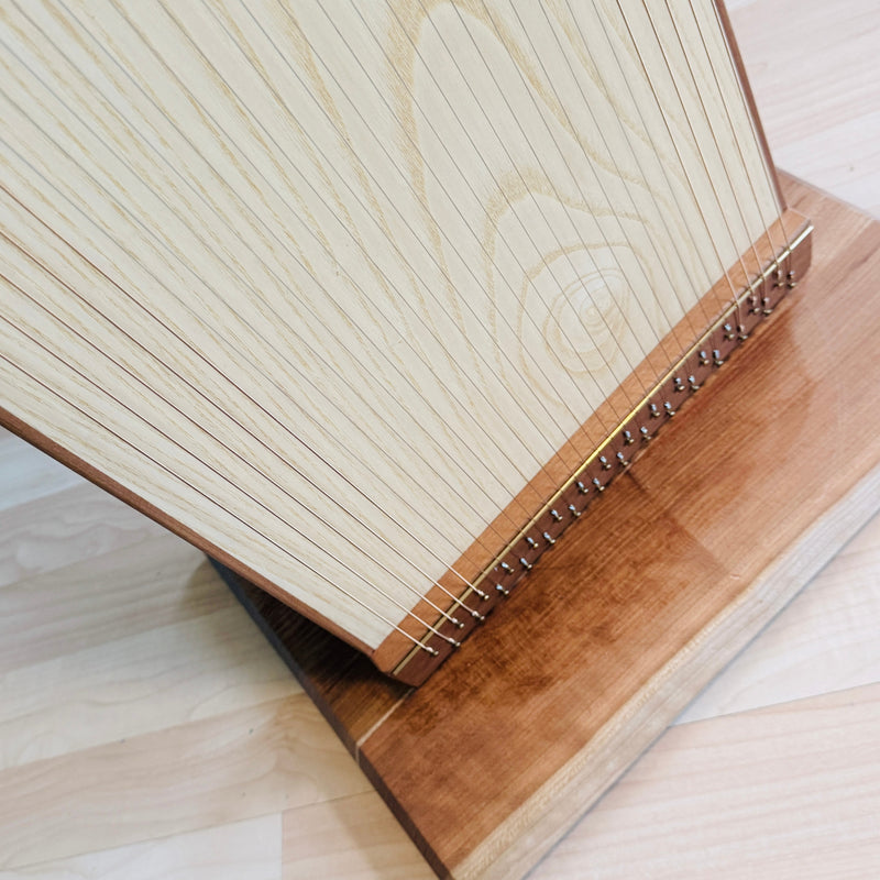 A close-up of a wooden stringed instrument, likely a zither or guitar-like plucking instrument, resting on a wooden stand. The instrument has a light natural wood body with many thin vertical strings and a darker brown wooden frame and base, photographed on a light wood floor.