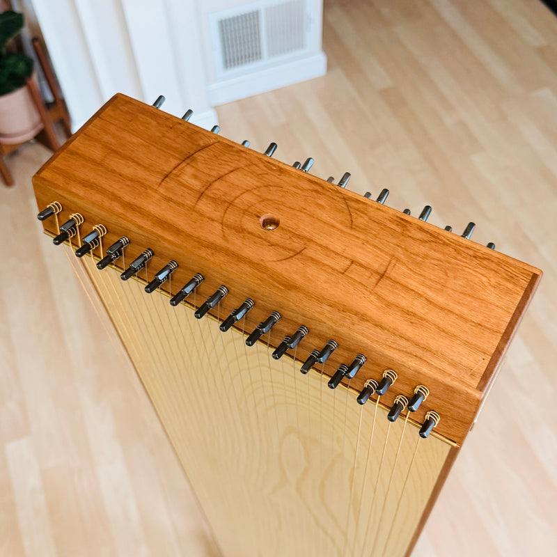 A close-up of a handcrafted wooden string instrument, likely a zither or guitar-like folk instrument, with a warm honey-toned wood top, light natural wood body, and black tuning pegs, photographed indoors on a light wood floor.