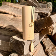 A natural Koshi Ocean  Earth Symphony Maple wind chime hanging outdoors from a tree trunk, with a small wooden tag below and a black hanging cord, set against a forest background.