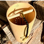 A natural Koshi Ocean Earth Symphony Maple wind chime hanging outdoors from a tree trunk, with a small wooden tag below and a black hanging cord, set against a forest background.