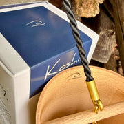 A natural Koshi Ocean Earth Symphony Maple wind chime hanging outdoors from a tree trunk, with a small wooden tag below and a black hanging cord, set against a forest background.