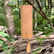 A natural Koshi River Earth Symphony  cherry wood wind chime hanging outdoors from a tree trunk, with a small wooden tag below and a black hanging cord, set against a forest background.