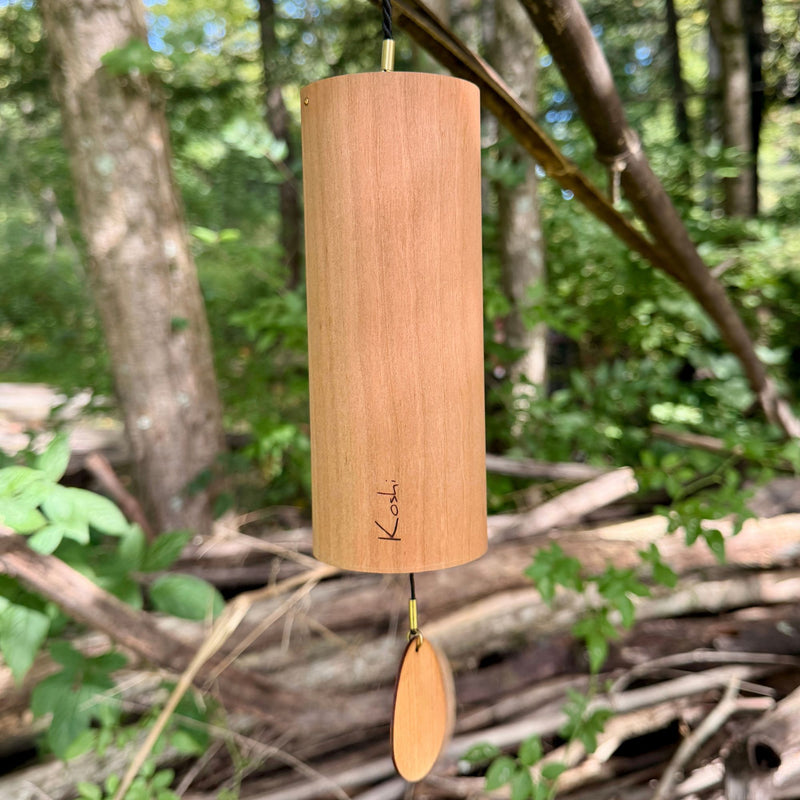 A natural Koshi River Earth Symphony  cherry wood wind chime hanging outdoors from a tree trunk, with a small wooden tag below and a black hanging cord, set against a forest background.