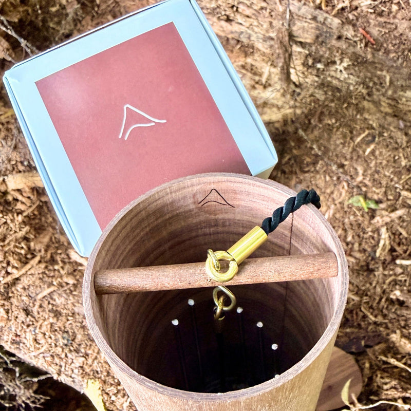 A natural Koshi Mountain Earth Symphony Walnut wind chime hanging outdoors from a tree trunk, with a small wooden tag below and a black hanging cord, set against a forest background.